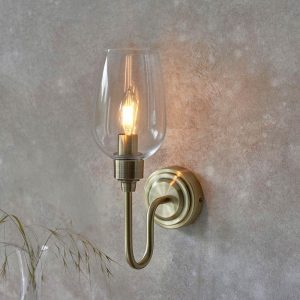 Classic antique brass wall light with clear glass shade, shown on wall in a room setting.
