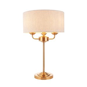 Highclere 3 light table lamp in vintage brass with natural linen shade, on white background, lit.