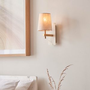 White marble wall light in vintage brass with natural linen tapered shade, shown on bedroom wall.