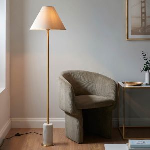 White marble designer floor lamp with tapered shade in aged brass, shown in a lounge setting.