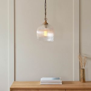 Ashcroft single light pendant in brushed aged brass with clear ribbed glass goblet shade, shown over living room table.