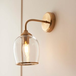 Ashcroft single wall light in brushed aged brass with clear glass goblet shade, shown on room wall.
