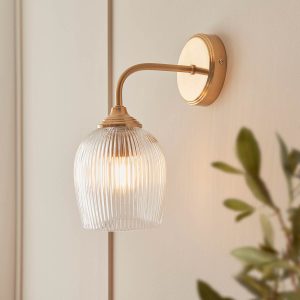 Ashcroft single wall light in brushed aged brass with clear ribbed glass goblet shade, shown on wall in a room setting.