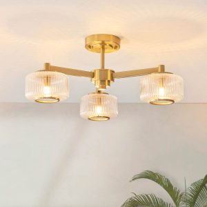 Stamford 3 light semi flush ceiling light in brushed aged brass with clear ribbed glass drum shades, shown on room ceiling.