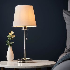 Polished nickel bedside table lamp with white shade, shown with vase on side table.