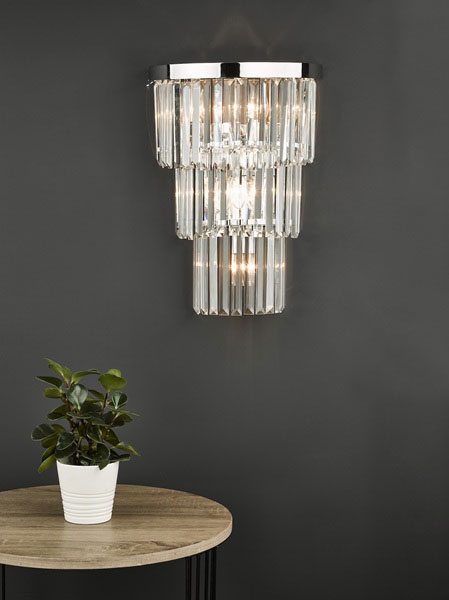 Angel chrome and crystal wall light shown lit on dark grey wall in room setting.