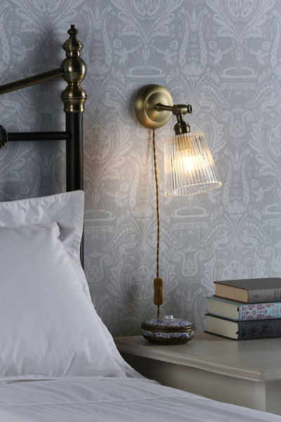 Laura Ashley Callaghan plug in wall light in antique brass, shown next to bed, lit.