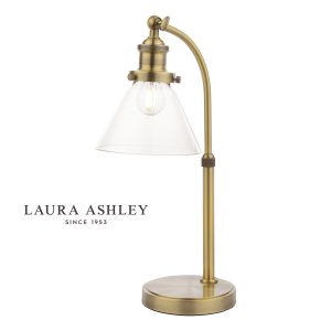 Laura Ashley Isaac desk lamp in antique brass finish with clear glass shade on white background, lit.