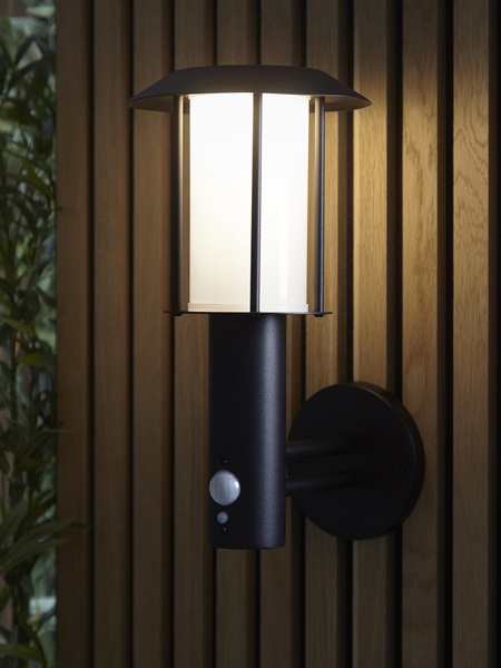 An illuminated traditional PIR lantern attached to a wooden garden fence at night